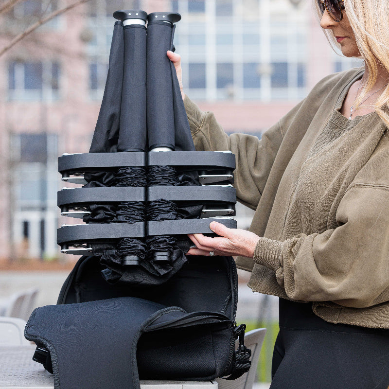 A person is setting up a black portable cot with an adjustable shoulder strap carrier in a park setting.