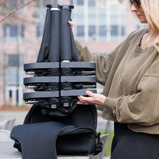 A person is setting up a black portable cot with an adjustable shoulder strap carrier in a park setting.